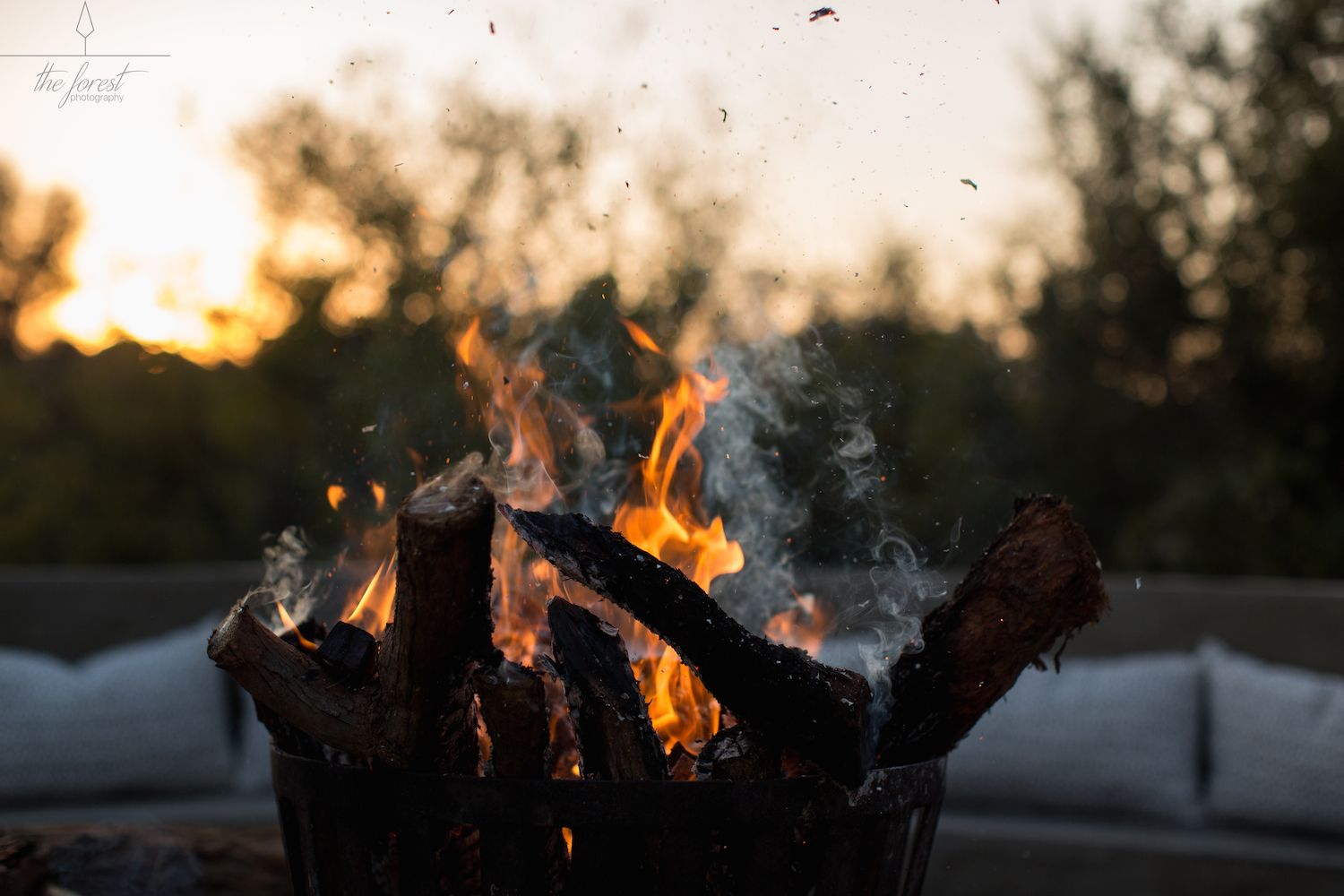 Een kampvuur met brandhout, omringd door een metalen bak, met op de achtergrond wazige bomen en een zonsondergang.