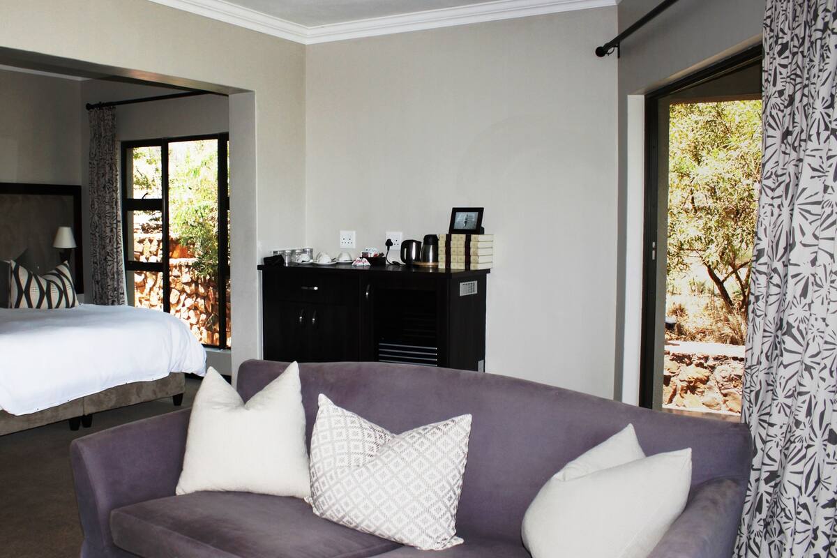 A bedroom with a bed, a sofa, a desk with a computer, and a window with a view of trees.