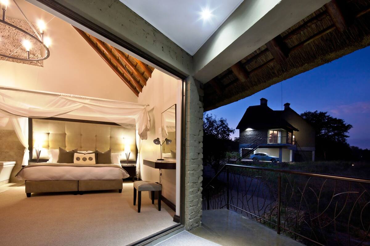 A bedroom with a large bed, bedside tables, and a nightstand. There is a window with a view of a house and a car outside.