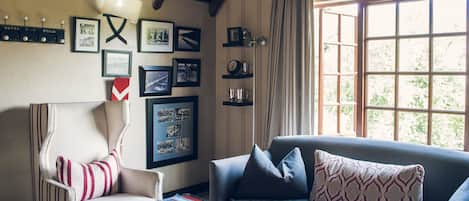 A living room with a sofa, armchair, and a table with magazines. There are framed pictures on the wall and a window with curtains.