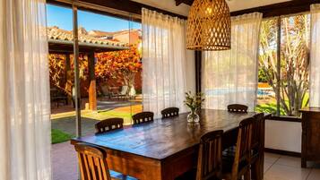 A wooden dining table with chairs, a hanging light fixture, and a view of a pool and garden through large windows.