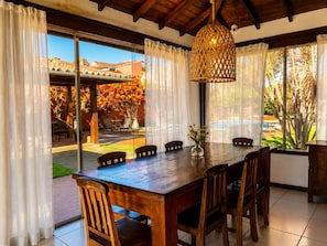 Una mesa de comedor de madera con sillas, una lámpara colgante y vistas a una piscina y jardín a través de amplios ventanales.