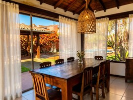 Un tavolo da pranzo in legno con sedie, un lampadario e vista su piscina e giardino attraverso ampie finestre.