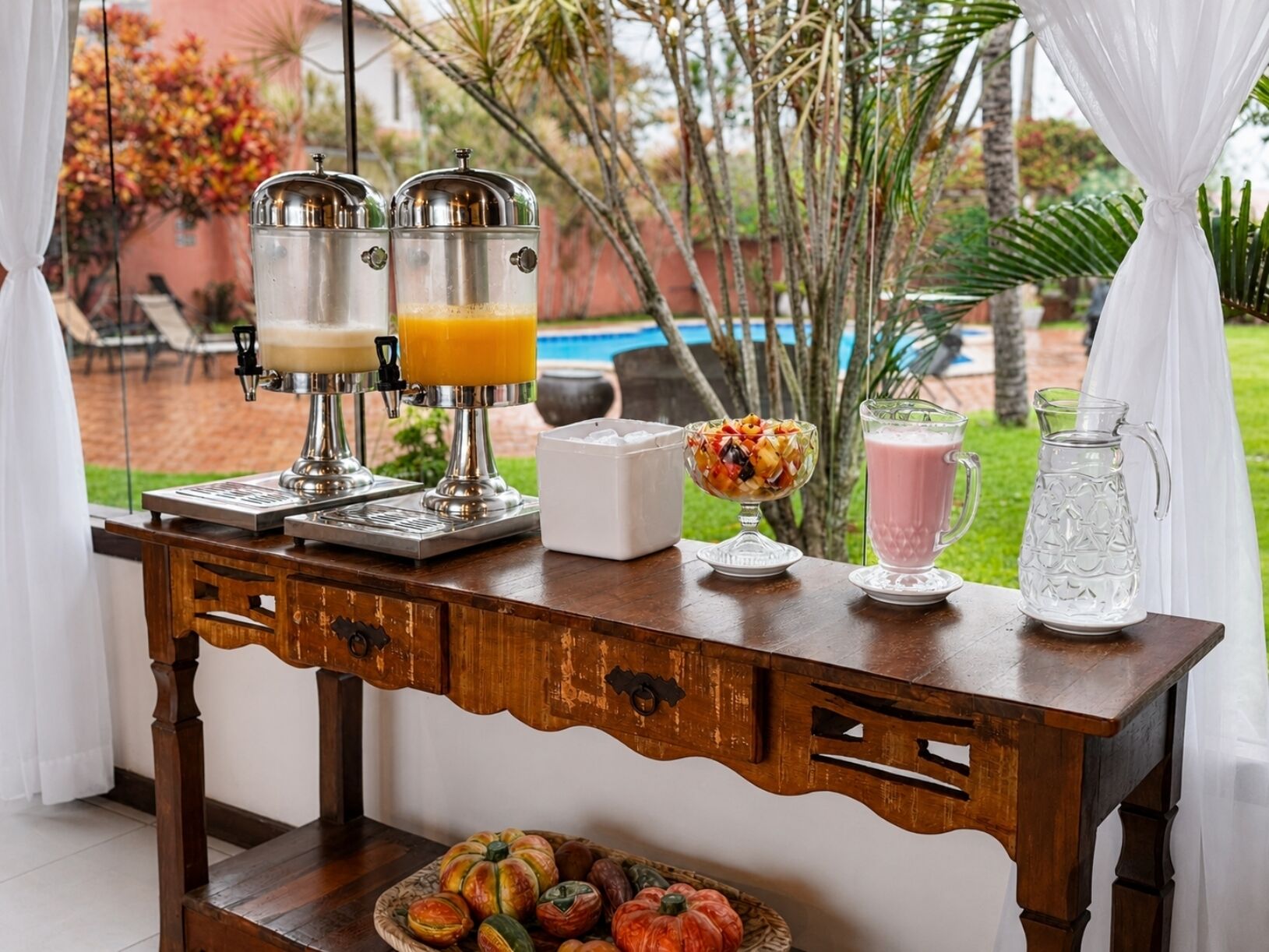 A wooden table with a coffee maker, a fruit bowl, a pitcher, and a glass pitcher.