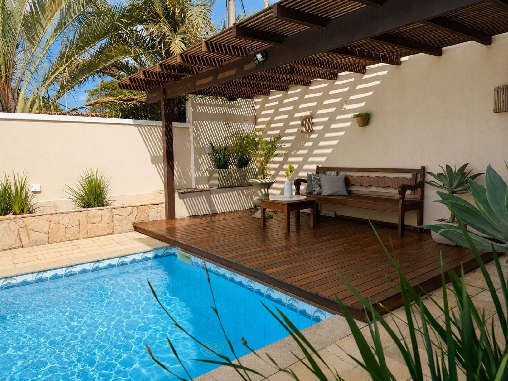 A swimming pool with a wooden deck, a bench, and potted plants.