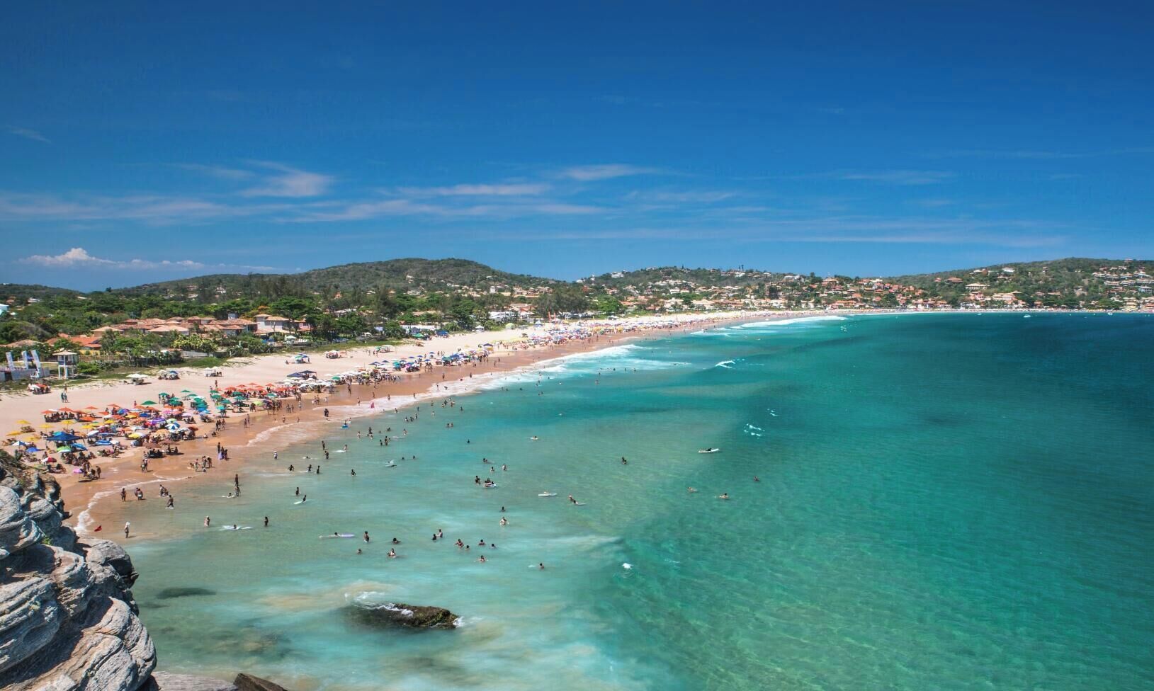 Uma praia com águas turquesa cristalinas, uma faixa de areia e um relevo acidentado ao fundo.