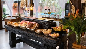A buffet table with various food items, a wooden bench, and a tropical plant. - Pousada Sable Dor (Búzios)