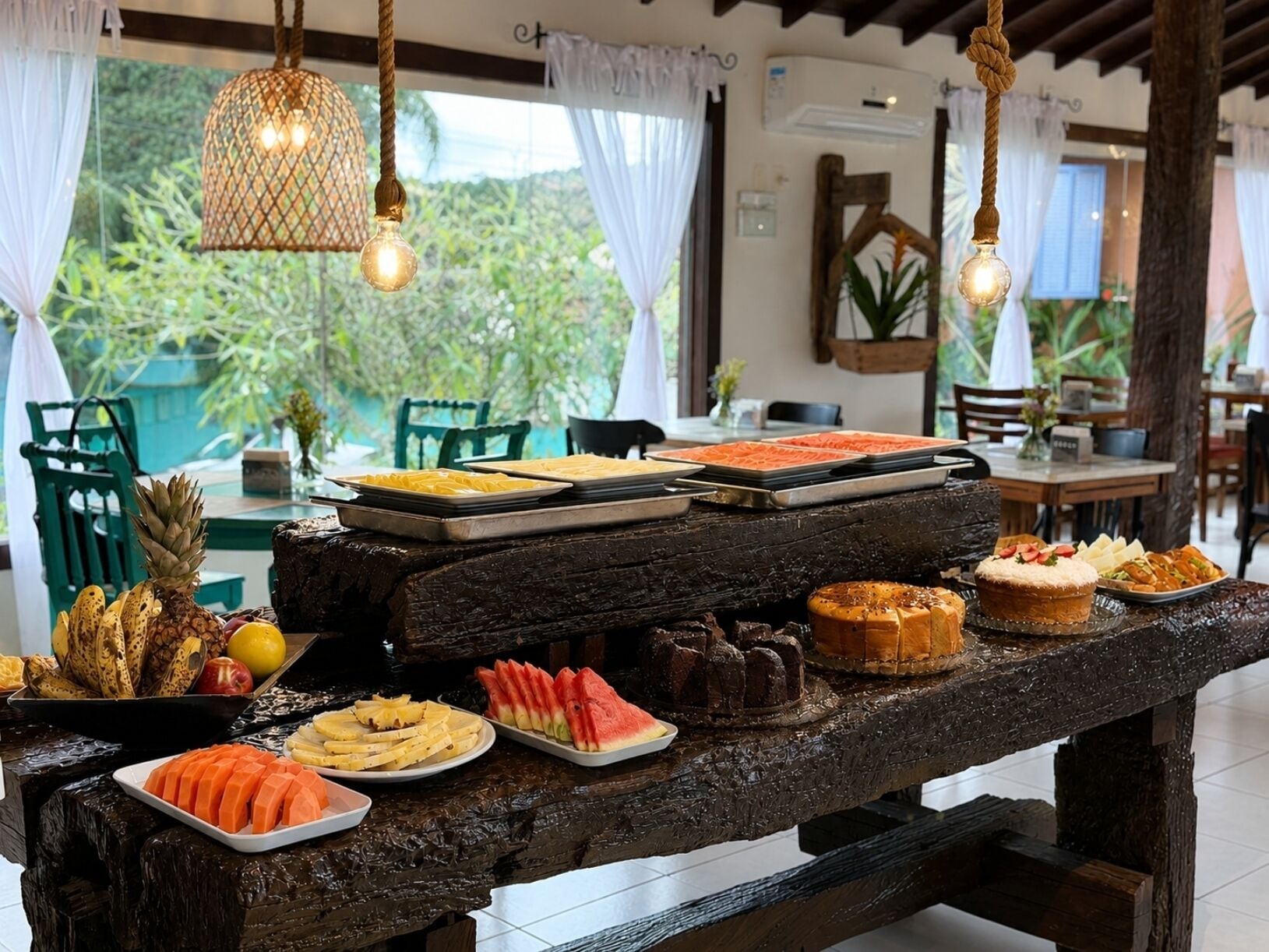 Uma mesa de madeira com diversas frutas e sobremesas, num espaço com luminárias pendentes e vista para o verde lá fora.