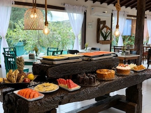 A wooden table with various fruits and desserts, set in a room with hanging lights and a view of greenery outside. - Pousada Sable Dor (Búzios)
