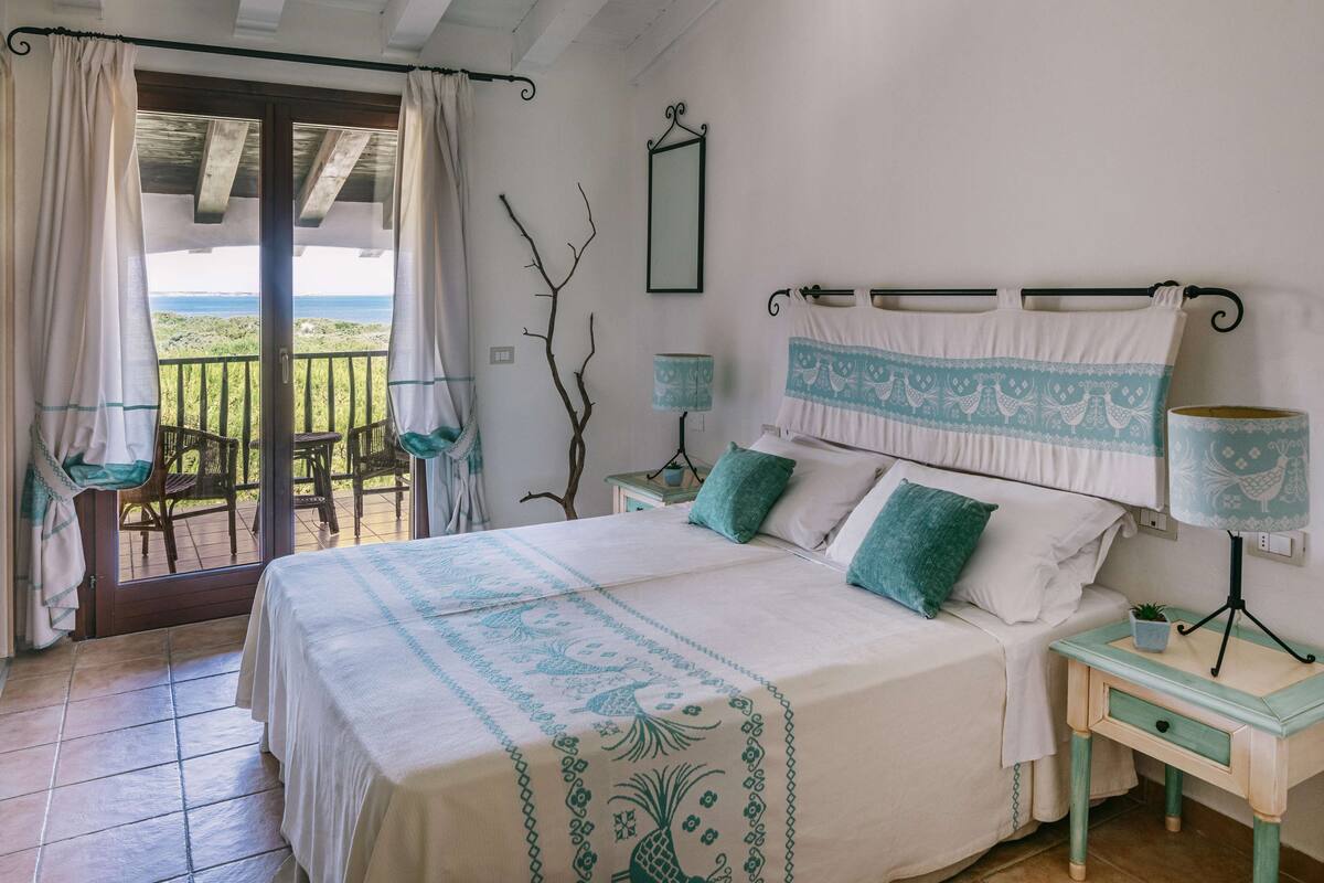 A bedroom with a bed, two bedside tables, a nightstand, a window with curtains, a view of the outdoors, and a wall-mounted mirror.