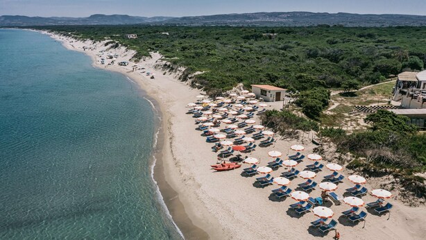 Une plage bordée de rangées de transats et de parasols, une mer d’un bleu limpide et un sentier menant à un bâtiment.