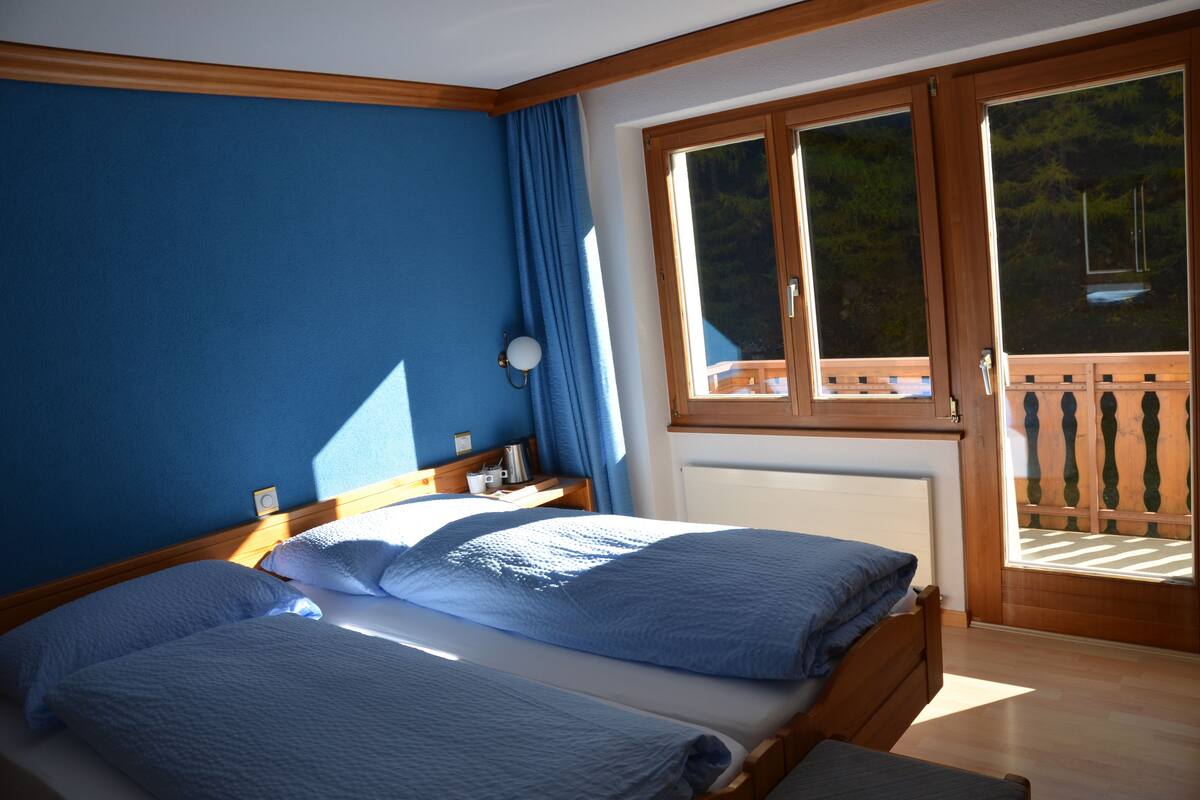 A bedroom with two beds, a wooden headboard, a nightstand, a lamp, and a window with a view of a balcony.