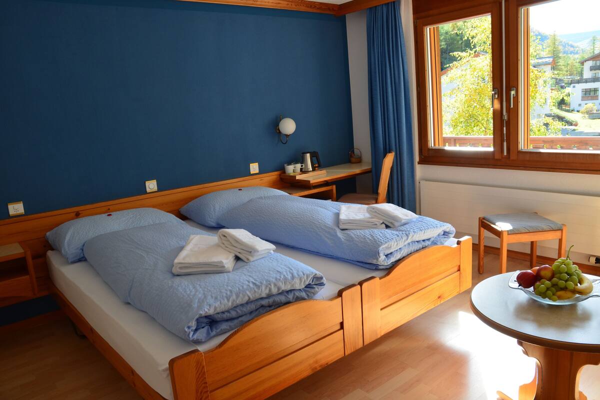 A double bed with blue bedding, a bedside table with a lamp, a small table with fruit, and a view of buildings through a window.