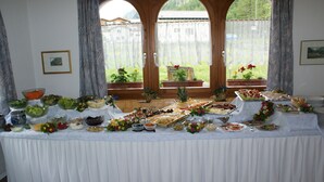 Una mesa de buffet con diversos alimentos y decoraciones, con vistas a ventanas con cortinas y plantas en macetas.