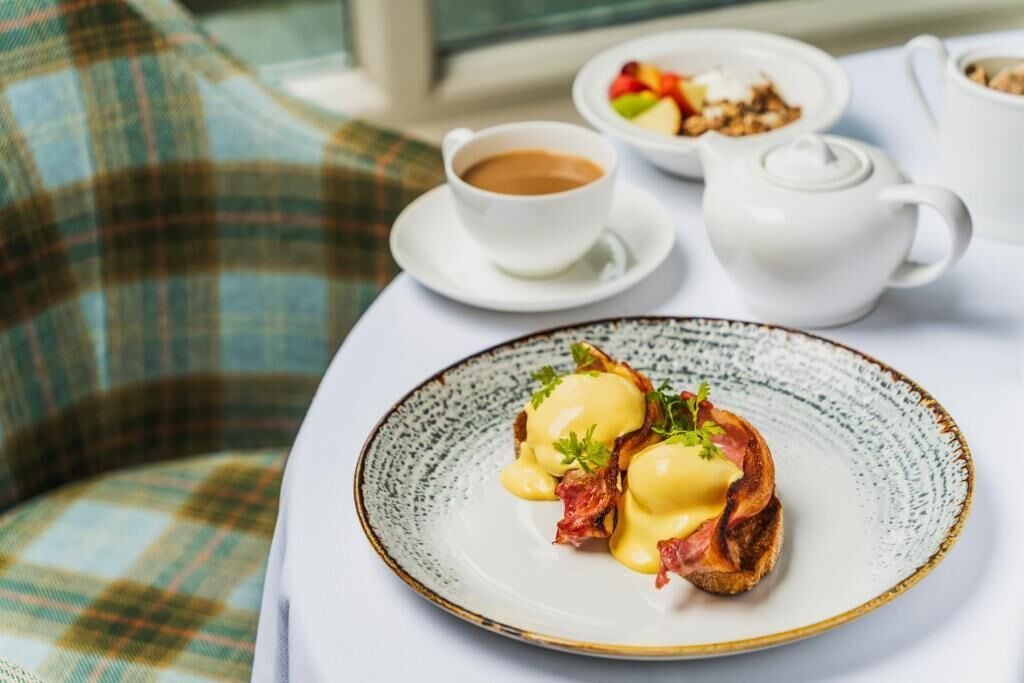 A breakfast setting with a plate of eggs benedict, a cup of coffee, and a bowl of fruit on a table.