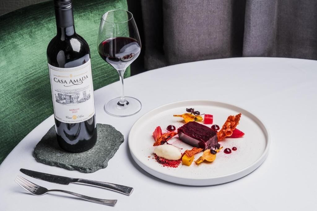 A bottle of Casa Amada Merlot 2023, a glass of red wine, and a plate of food on a white table with a green chair in the background.