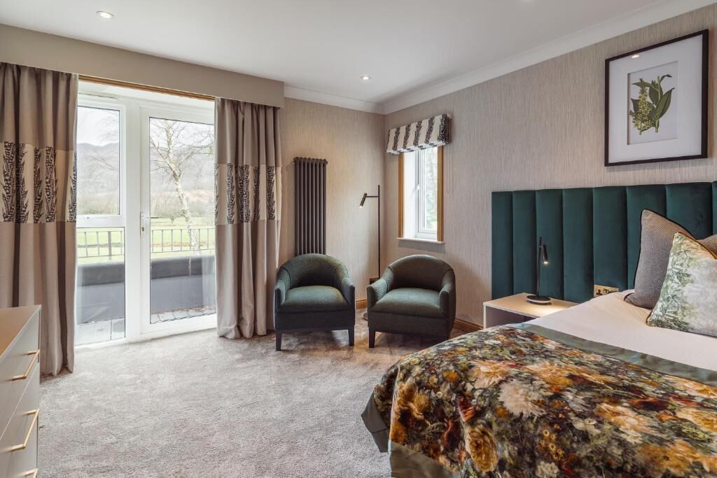A bedroom with a bed, two armchairs, a dresser, a window with curtains, and a framed picture on the wall.