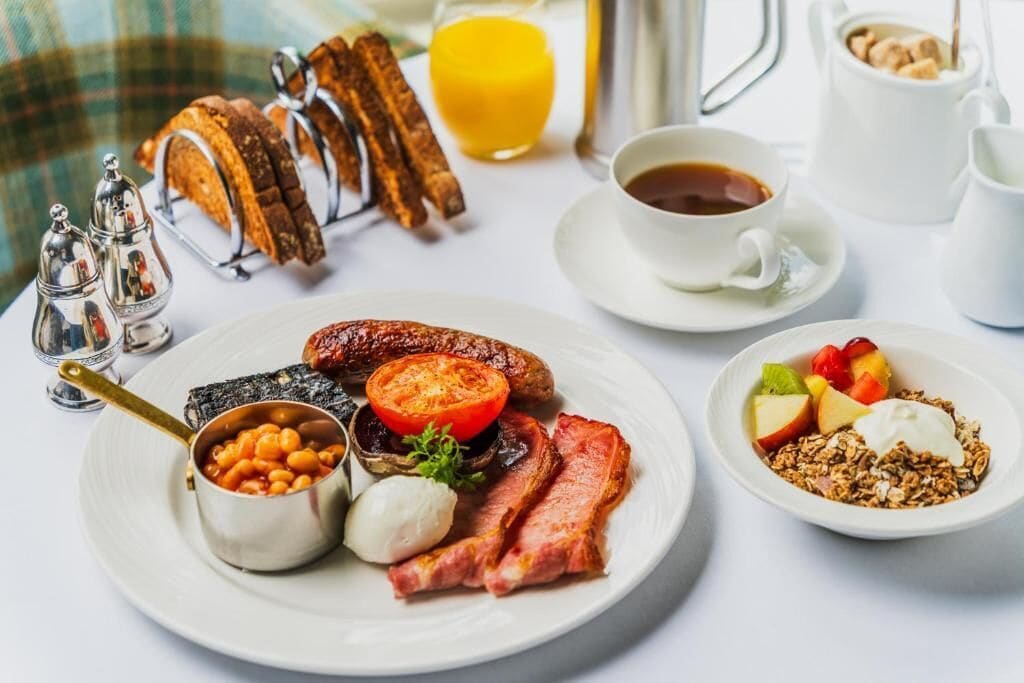 A breakfast setting with a plate of eggs, bacon, sausages, beans, and tomatoes, a bowl of granola with fruit, and a cup of coffee.