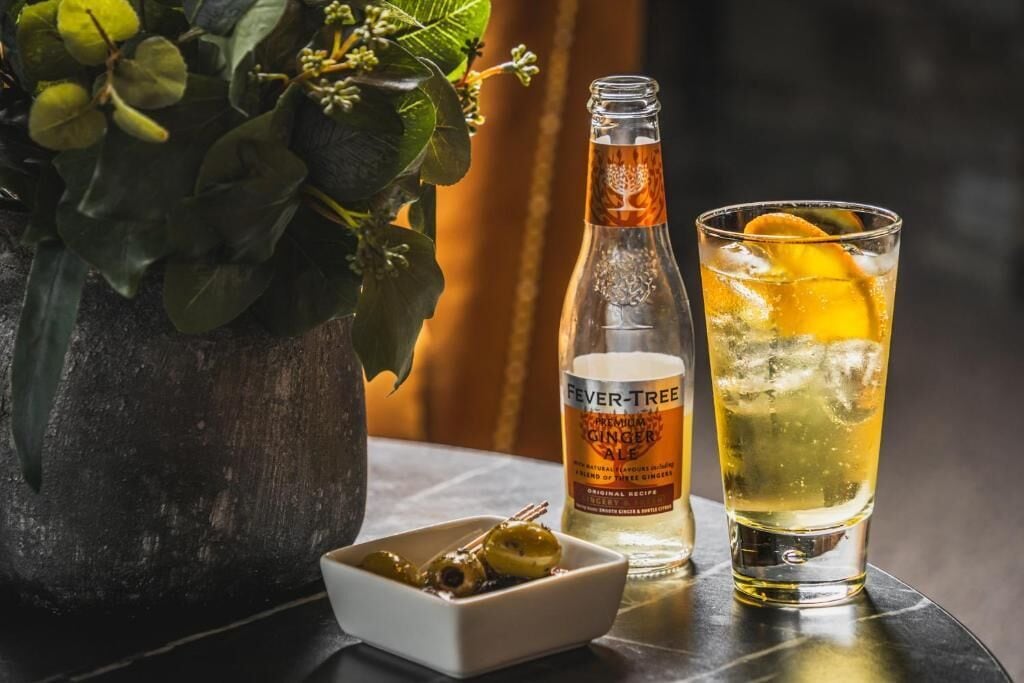 A bottle of Fever-Tree Ginger Ale and a glass with ice and an orange slice on a table.
