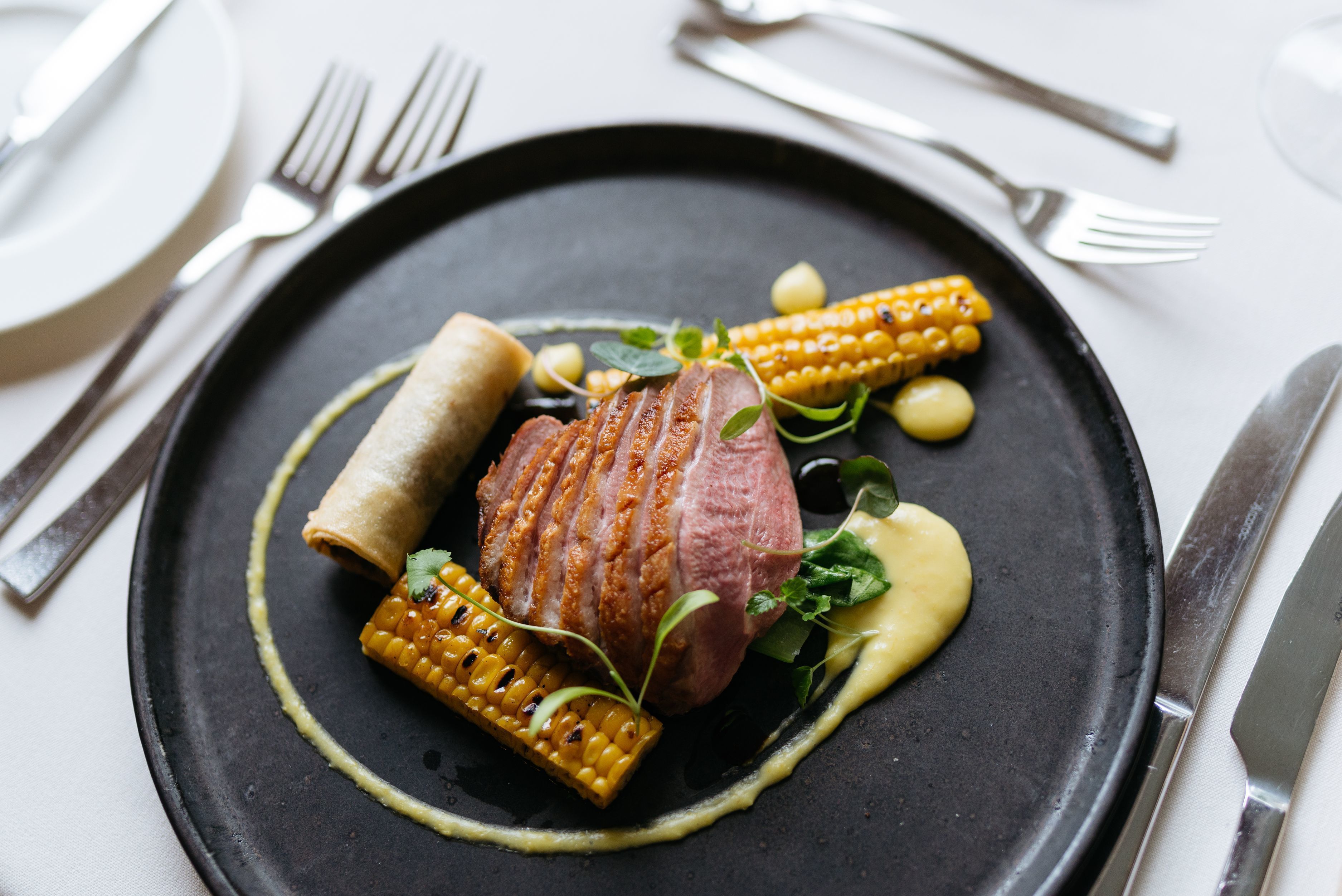 A black plate with a piece of meat, corn on the cob, and a small roll.