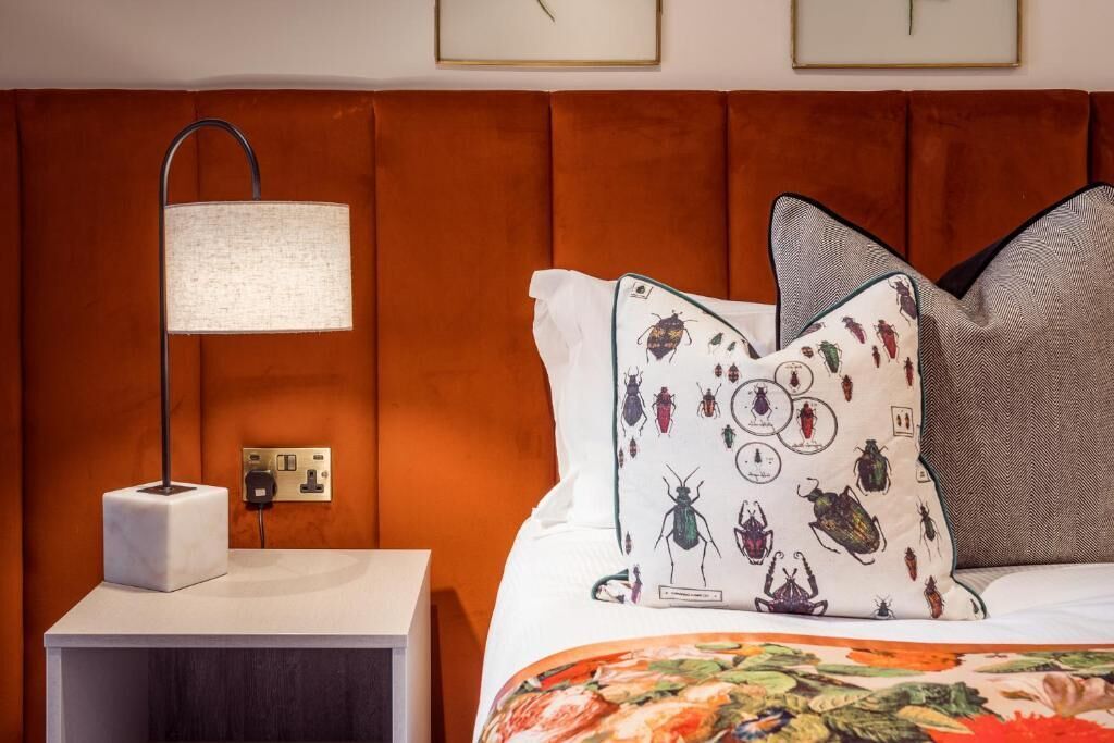 A hotel room with a brown headboard, a bedside lamp, a white nightstand, and a bed with a floral bedspread and patterned pillows.
