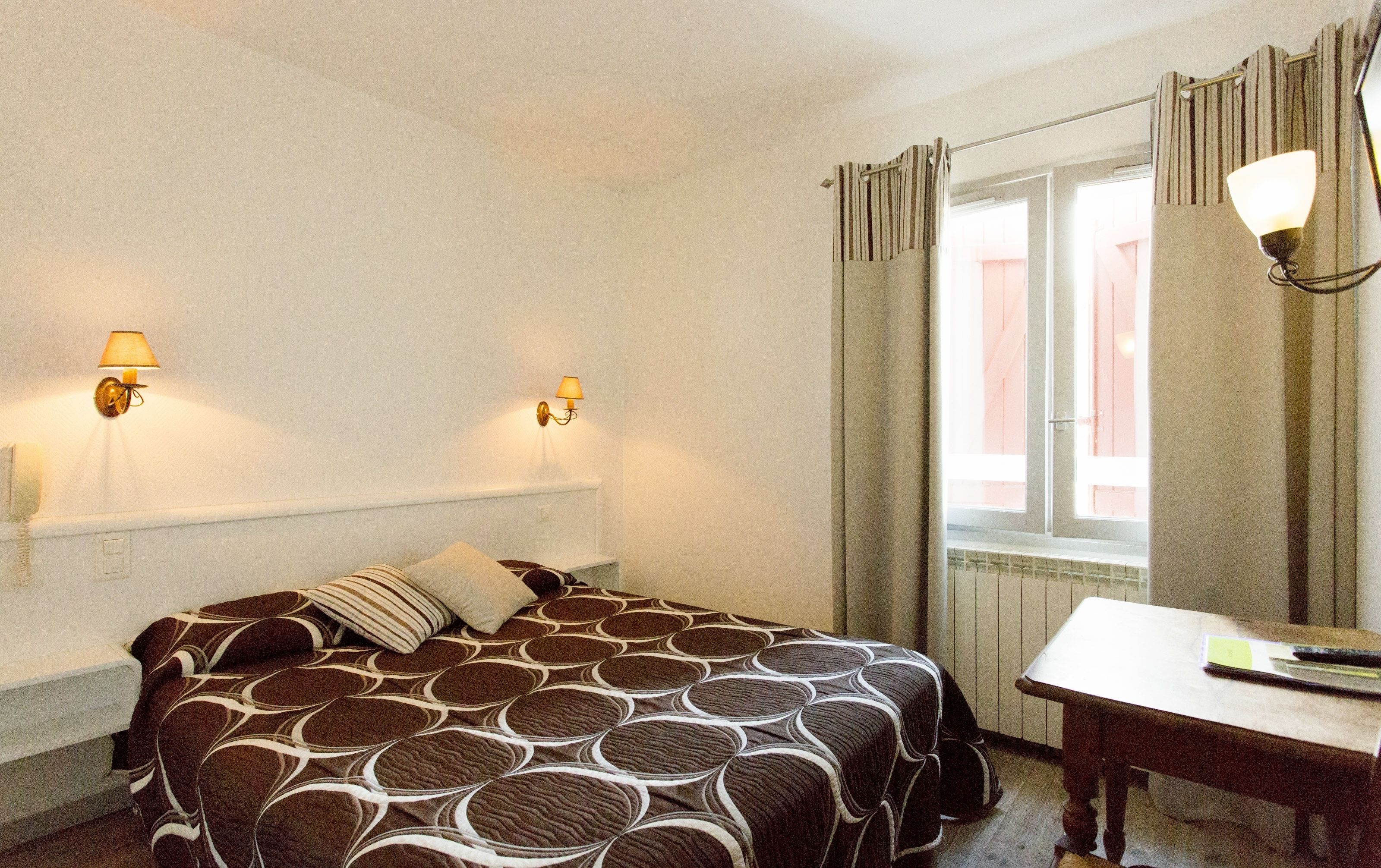 A hotel room with a bed, two wall-mounted lamps, a window with curtains, a small table, and a chair.