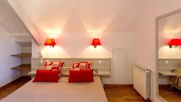 Ein Schlafzimmer mit schrägem Dach, zwei Nachttischlampen, einem Bett mit rot-beiger Bettwäsche und einem großen Spiegel an der Wand.