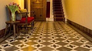 A hallway with a checkered floor, a wooden door, a staircase, and a chair with a vase of flowers. - Hôtel Etchémaïté (Larrau)