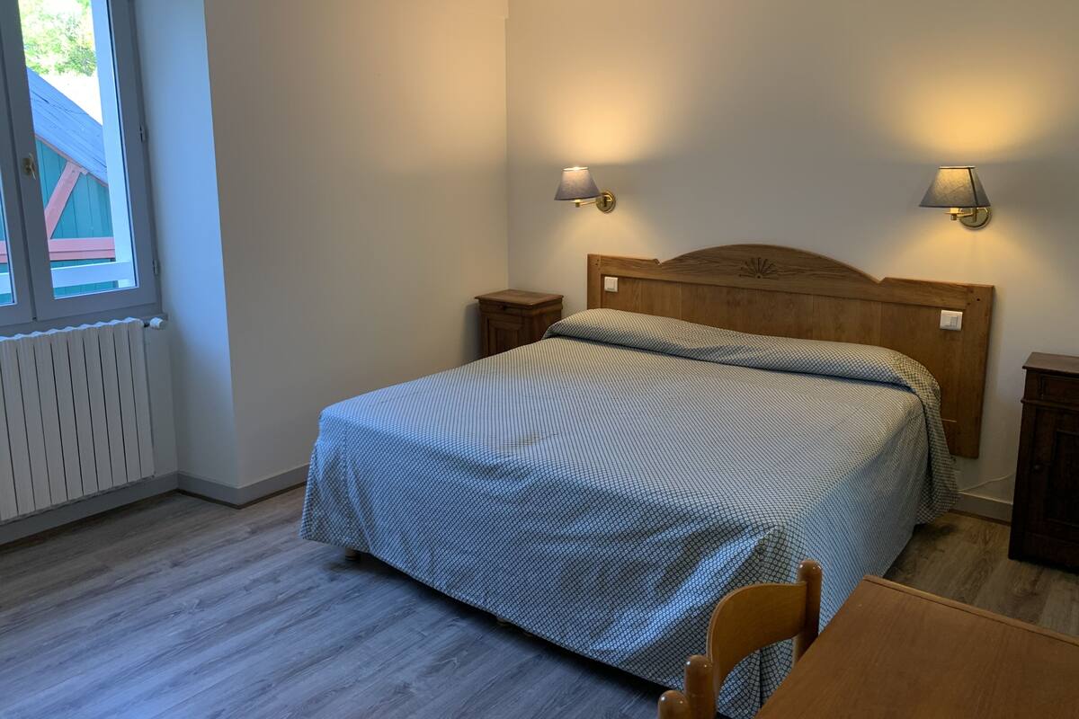 Ett sovrum med en säng, två sängbord, ett litet bord, en träpanel vid sänggaveln, ett fönster och ett element.