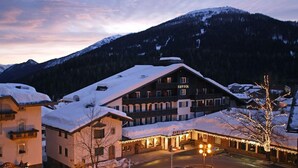 Un albergo con tetto imbiancato dalla neve, un cartello con su scritto "SAVOIA" e montagne sullo sfondo.