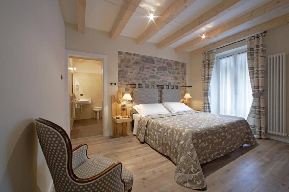 Een slaapkamer met een bed, een stoel, een stenen muur, een raam met gordijnen en een badkamer in de achtergrond.