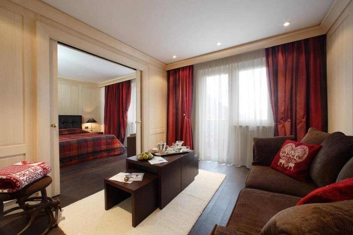 Een moderne hotelkamer met een groot bed, een zithoek met een bank en fauteuil, een klein bureau en een groot raam met gordijnen.
