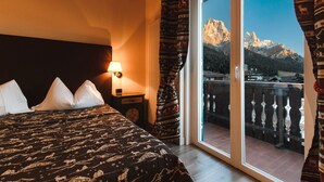 Una camera d'albergo con un letto, un comodino, una lampada e vista sulle montagne da un balcone.