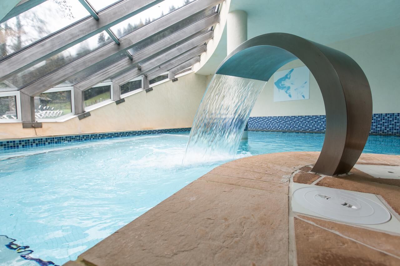 Una piscina interior con caída de agua, techo de cristal y borde de azulejos.