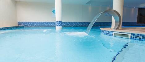 An indoor swimming pool with clear blue water, a central water feature, and a tiled border.