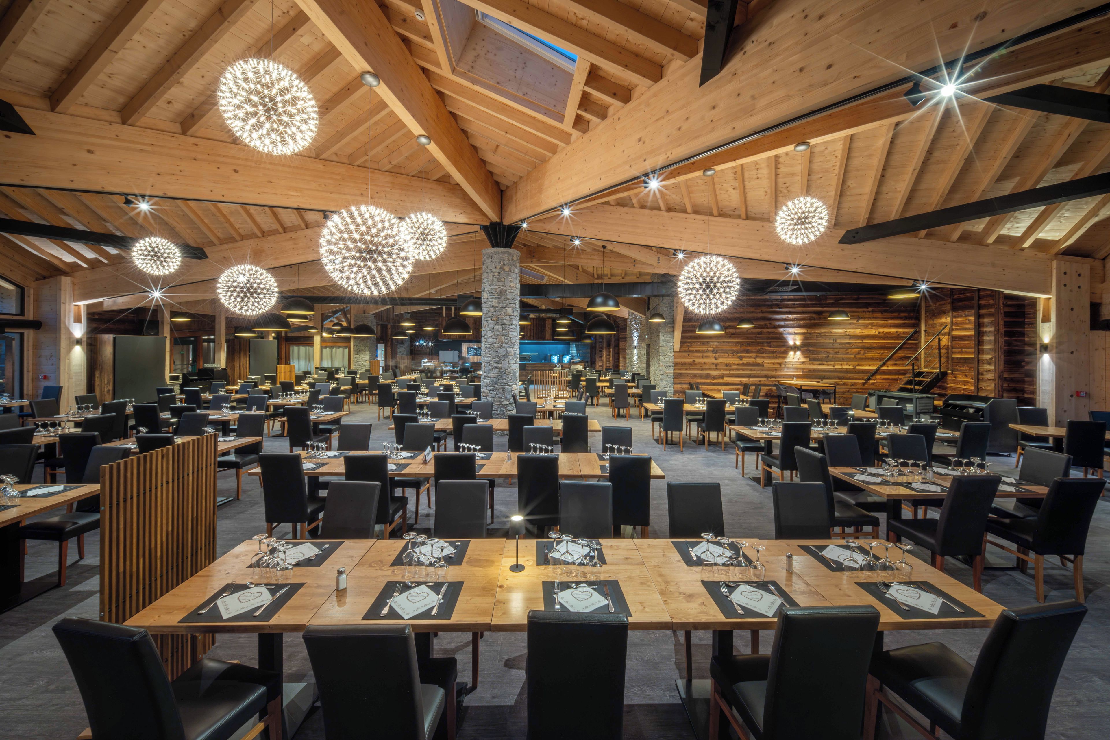A spacious dining area with multiple tables and chairs, wooden beams on the ceiling, and hanging spherical light fixtures.