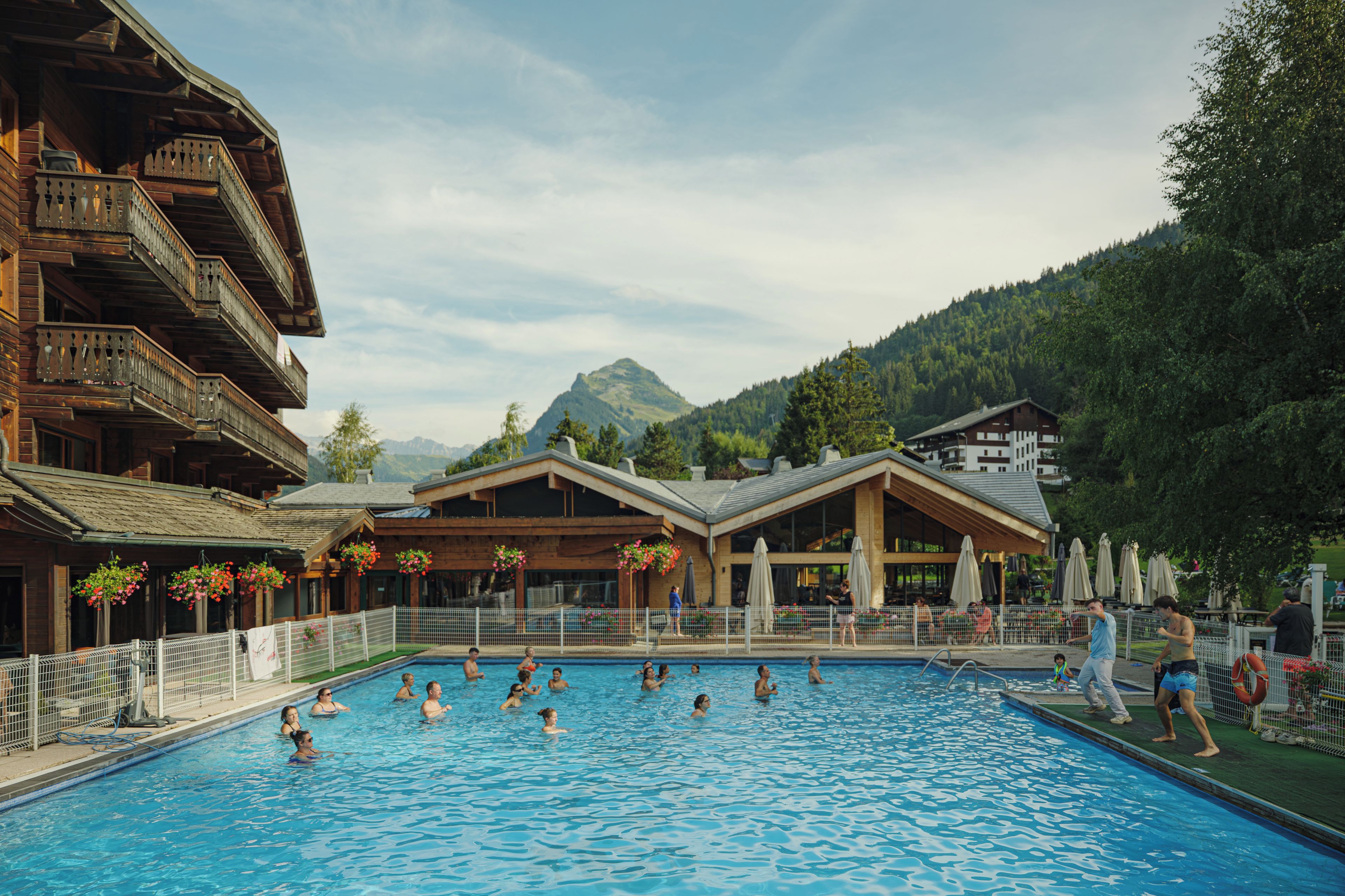 A swimming pool surrounded by wooden buildings with balconies, set against a mountainous backdrop.