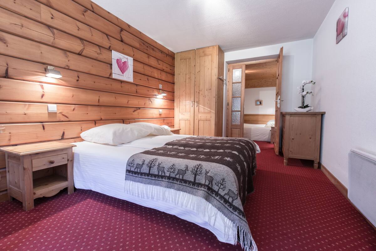 A bedroom with a wooden headboard, a bed with a patterned blanket, a nightstand, a wooden wardrobe, and a red carpeted floor.