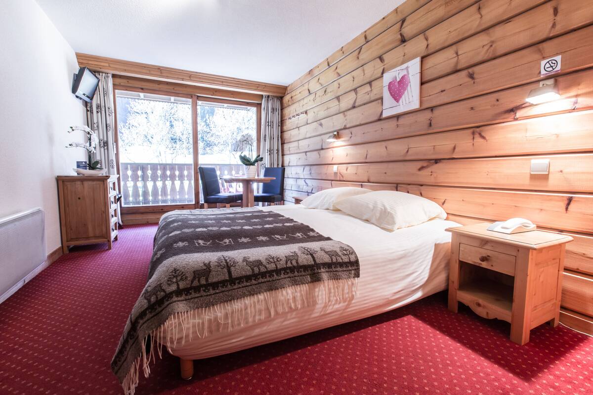 A bedroom with a bed, wooden headboard, a nightstand, a small table with chairs, and a window with a view of trees.