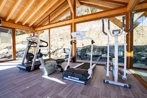 A gym with treadmills, elliptical machines, and a weight machine, featuring large windows and wooden beams. - Hôtel Club le Crêt (Morzine)