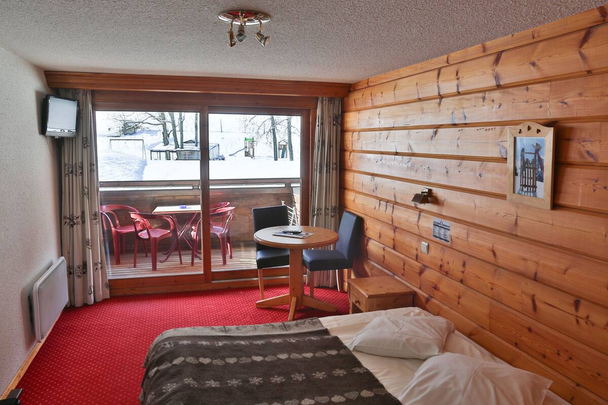 A room with a bed, a small table with chairs, a wooden wall with a picture, and a view of the outside.