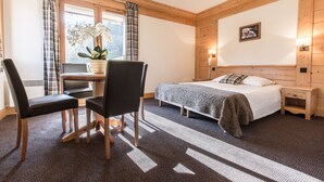 Ein Hotelzimmer mit Bett, Schreibtisch mit zwei Stühlen, Fenster mit Vorhängen, Nachttisch mit Lampe und einem an der Wand befestigten Telefon.