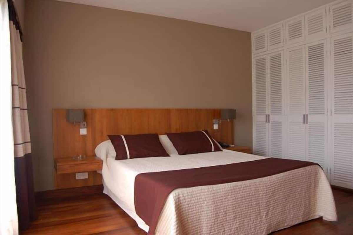 A bedroom with a wooden headboard, a bed with a white and brown bedspread, two bedside tables, and a wardrobe with louvered doors.