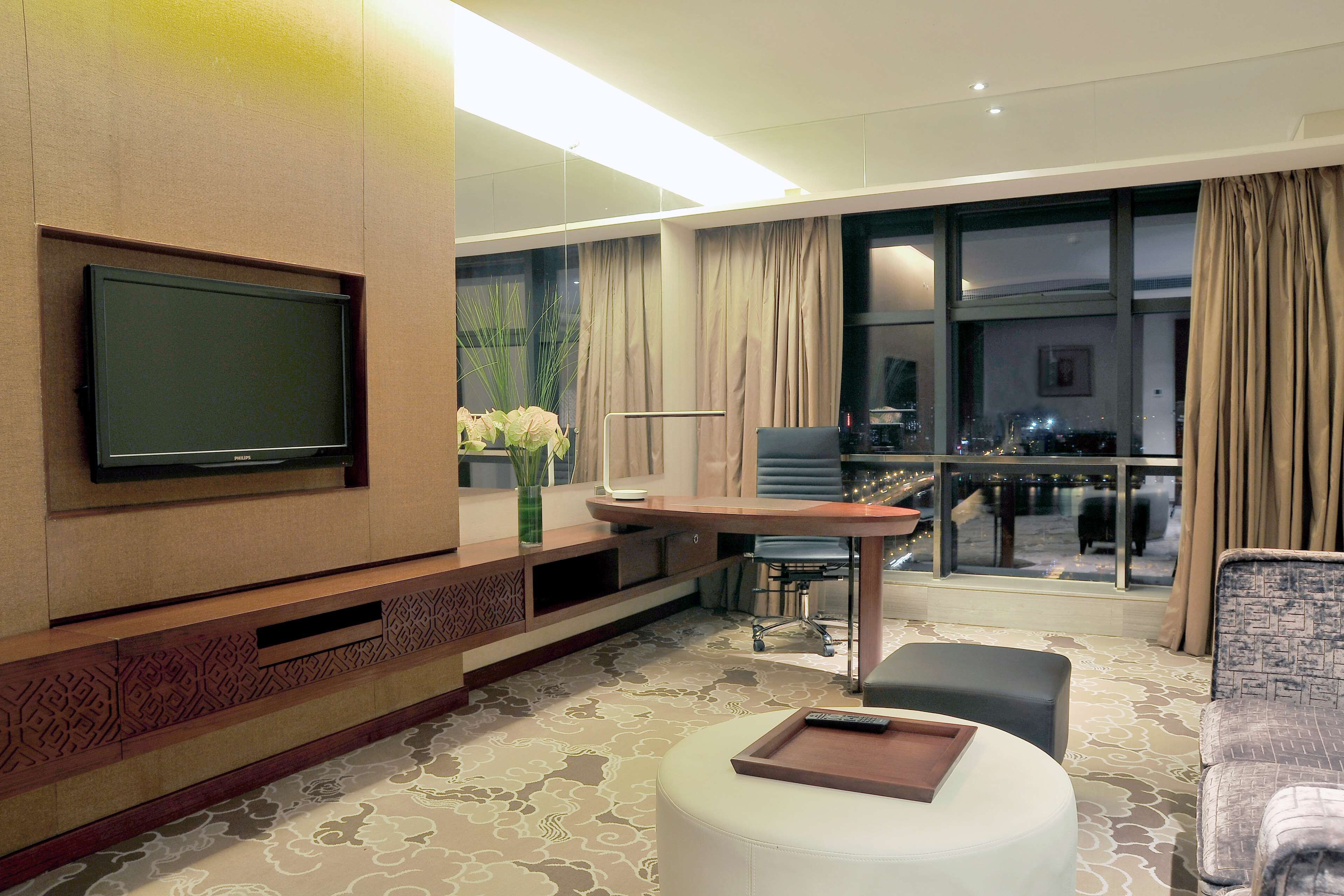 Una camera d'albergo moderna con una TV a schermo piatto montata a parete, un'area pranzo con tavolo e sedie, e un'ampia finestra con tende.