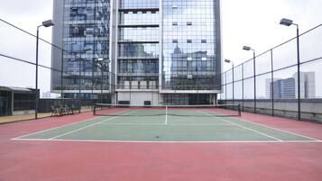 An outdoor tennis court with a net in the center, surrounded by a tall building with multiple floors and large windows.