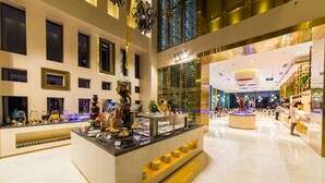 Um lobby de hotel moderno com área central de alimentação, balcão de recepção e um espaço amplo, com pé-direito alto e janelas grandes.