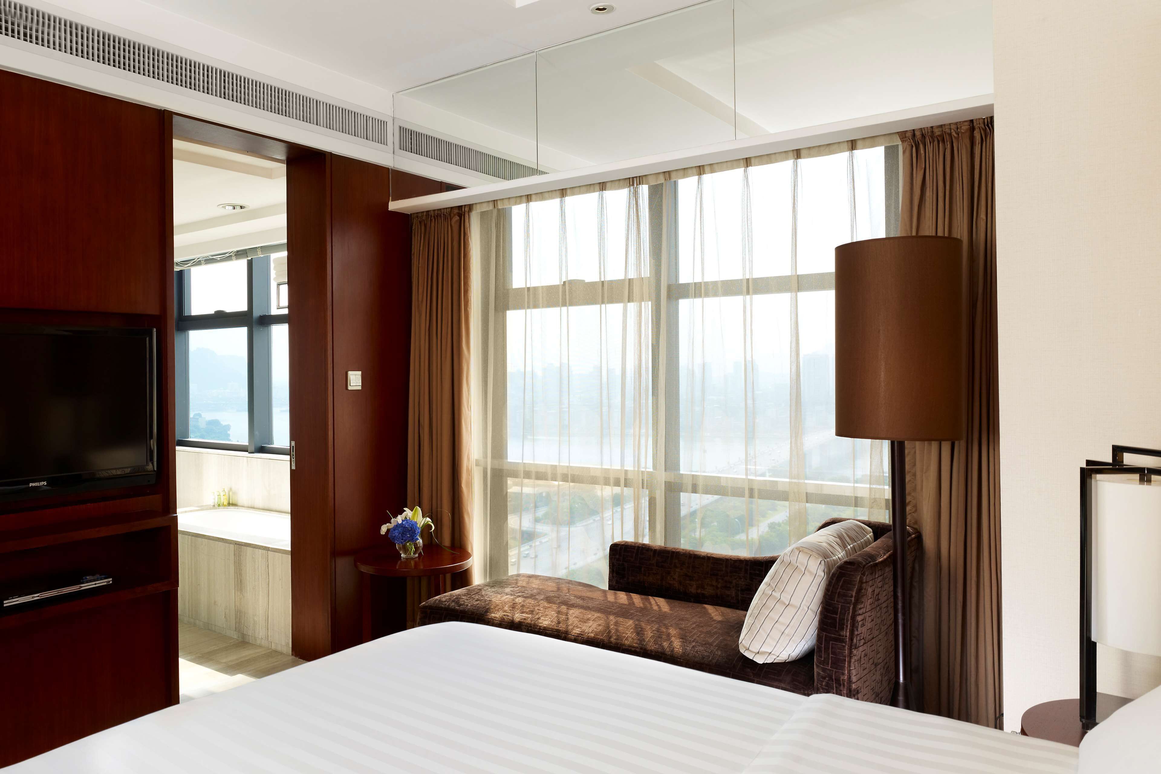 Ein Hotelzimmer mit einem großen Bett, einem Fernseher, einer Sitzecke mit einem Sessel, einem Tisch mit einer Vase und Blick auf die Stadt durch große Fenster.