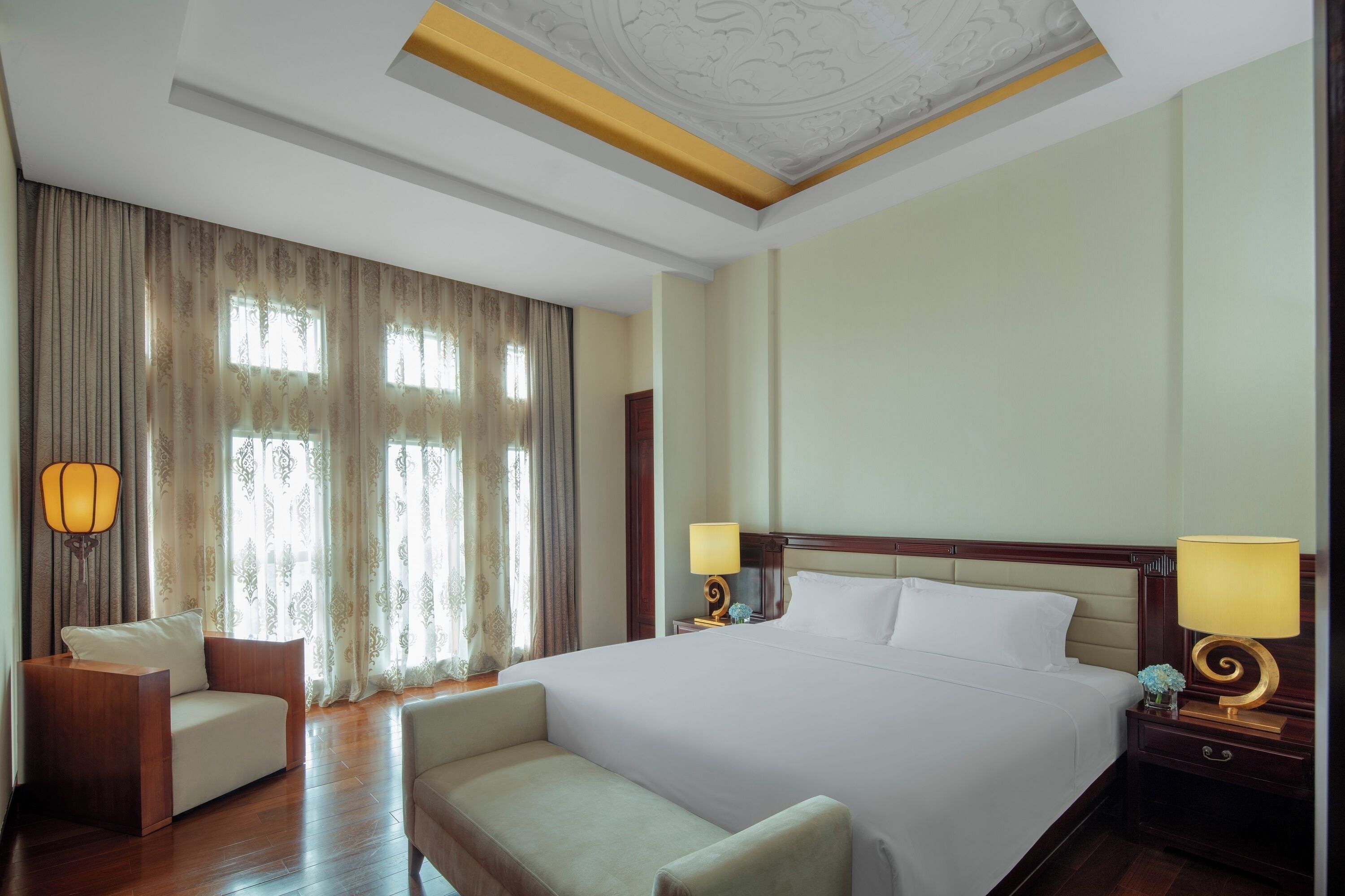 Habitación de hotel con una cama grande, dos mesitas de noche con lámparas, un sofá y un amplio ventanal con cortinas.