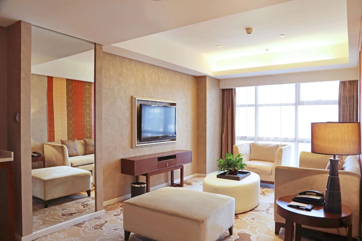 Ein Hotelzimmer mit einem großen Flachbildfernseher an der Wand, einem Sofa, einem Hocker, einem kleinen Tisch mit einer Lampe und einem großen Fenster, das natürliches Licht hereinlässt.