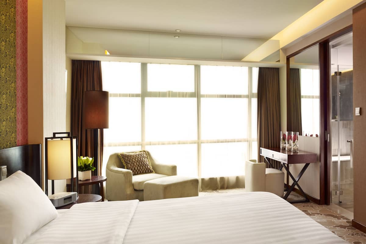 Ein Hotelzimmer mit einem großen Bett, einem Schreibtisch mit Stuhl, einem an der Wand befestigten Fernseher und einem großen Fenster mit Vorhängen.
